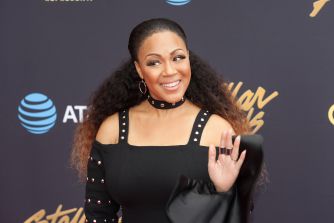 Erica Campbell smiles and waves at an event while wearing a black outfit.