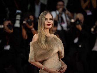 Angelina Jolie poses on a red carpet in a fur-trimmed dress as photographers take pictures behind her.