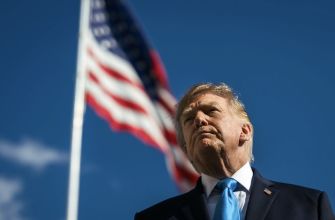 President Donald Trump stands outdoors in a suit and blue tie with an American flag behind him.