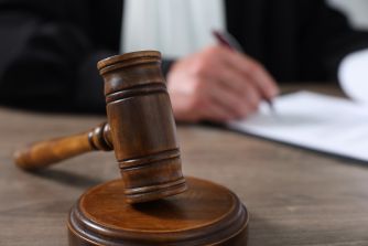 A wooden judge’s gavel rests on its sound block in a courtroom, with a blurred person writing documents in the background.