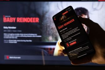 A hand holds a smartphone displaying Netflix’s Baby Reindeer page in front of a television showing the series title.