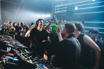 Sara Landry performs at a DJ console while people gather around her in a crowded club.