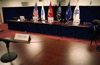Empty military commission courtroom with a prosecution table, microphones, and U.S. and military flags.