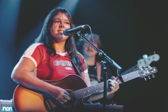 Gigi Perez performs onstage, singing into a microphone while playing an acoustic guitar.