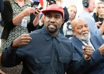Kanye West, wearing a red cap and dark shirt, gestures while standing among people at a public event.