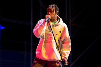 Travis Scott performs into a microphone onstage, wearing a white graphic hoodie.
