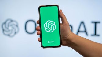 Hand holding a smartphone displaying the OpenAI logo in front of a blurred OpenAI sign.