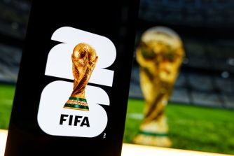FIFA World Cup trophy logo displayed on a black sign beside a blurred golden trophy on a soccer field.