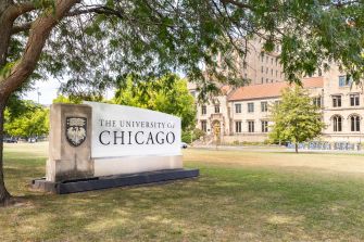 University of Chicago sign and campus buildings viewed from a grassy lawn.