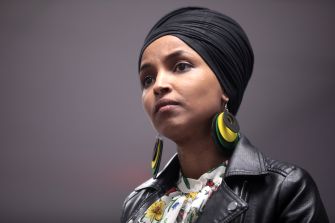 Rep. Ilhan Omar wearing a black headwrap, leather jacket, and large earrings while looking ahead.