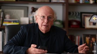 Larry David sits at a desk holding a phone and gesturing in front of bookshelves.