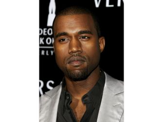 Kanye West poses at a red-carpet event wearing a gray blazer over a black shirt.