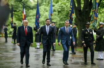 Donald Trump walks alongside military officials on a rain-soaked path lined with American and military flags.