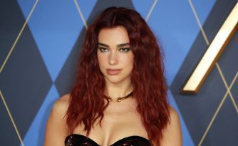 Dua Lipa poses with long red hair against a geometric blue backdrop.