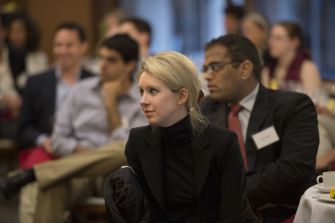 Elizabeth Holmes sits among attendees at a conference, wearing a black turtleneck and blazer.