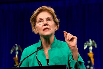 Sen. Elizabeth Warren speaks at a podium, raising one hand against a blue curtain backdrop.