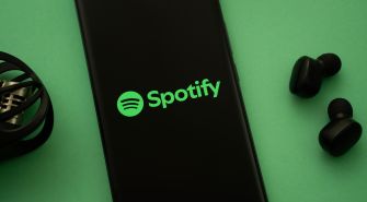 Spotify logo displayed on a smartphone beside wireless earbuds on a green surface.