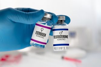 Gloved hand holding vials labeled estrogen and testosterone hormones.