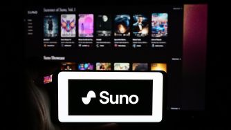 Suno logo displayed on a tablet in front of a computer screen showing the Suno music-generation interface.