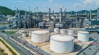 Oil refinery complex with storage tanks, processing towers, pipelines and surrounding hills.