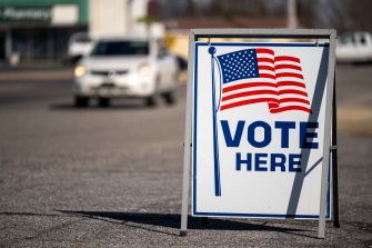 “VOTE HERE” sign displayed on a roadside near a polling place, with cars passing in the background.