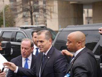 A suited man gestures toward reporters while walking beside several men and black SUVs.