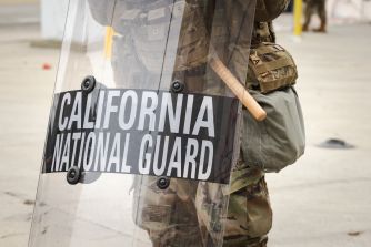 California National Guard member in riot gear holding a shield labeled “California National Guard.”
