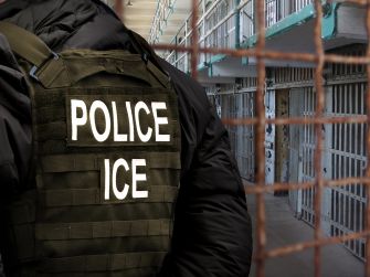 ICE officer wearing a vest labeled “POLICE ICE” stands outside barred detention facility cells.