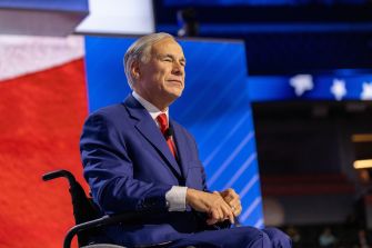 Texas Gov. Greg Abbott sits in a wheelchair at an event, wearing a blue suit and red tie.