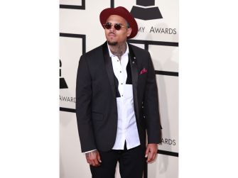 Chris Brown poses on the red carpet in a black suit, white shirt, burgundy hat, and sunglasses.