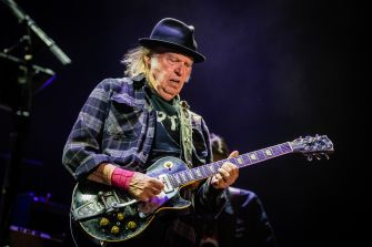 Neil Young performs on electric guitar under stage lights, wearing a black hat and plaid shirt.