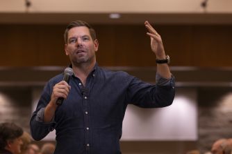 Eric Swalwell speaks into a microphone while raising one hand during an indoor event.
