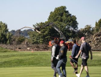 Workers walk across a golf course as an excavator removes trees and brush in the background.
