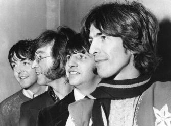Four Beatles members stand closely together, smiling in a black-and-white portrait.
