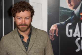 Danny Masterson stands at an event wearing a textured blazer, with a promotional poster visible behind him.