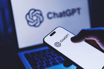 A hand holds a smartphone displaying the ChatGPT logo in front of a laptop showing the ChatGPT website.