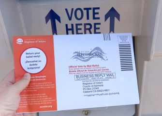 A hand holds a California vote-by-mail return envelope in front of a “VOTE HERE” sign.