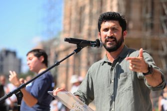 Man speaking into a microphone and gesturing while holding papers at an outdoor event.