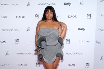 A woman poses on a step-and-repeat backdrop wearing a gray off-the-shoulder, ruffled mini dress.