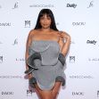 A woman poses on a step-and-repeat backdrop wearing a gray off-the-shoulder, ruffled mini dress.