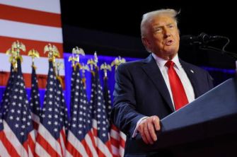 Donald Trump speaking at a podium with American flags in the background.