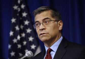 Image of Xavier Becerra speaking at a podium with a microphone, in front of an American flag backdrop.