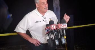 Male official in a white polo shirt speaks at a nighttime press conference, with yellow police tape and multiple microphones in front.