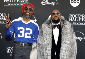 Two men posing on a Rock Hall red carpet, one in a blue jersey with a red beanie and the other in a fur coat with sunglasses, with Disney+ and Rock Hall logos in the background.
