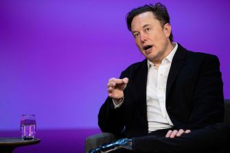 Elon Musk speaking during an event, with a glass of water on a table beside him.