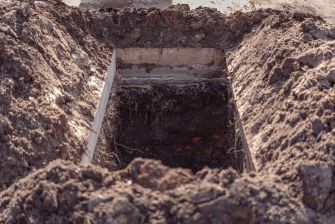 Deep rectangular trench in brown soil with wooden edging and visible roots.