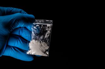 A gloved hand holding a small plastic bag containing a white powder, potentially indicating a dangerous drug.