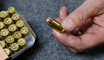 A close-up of a hand holding a bullet, with a box of ammunition in the background.