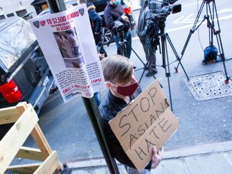 A person holding a handmade sign that reads "STOP ASIAN HATE" stands beside a poster offering a reward for information about an anti-Asian hate crime.