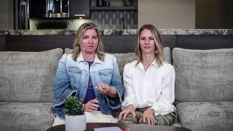 Two women sitting on a couch, discussing a topic, with a plant and papers on the table in front of them.